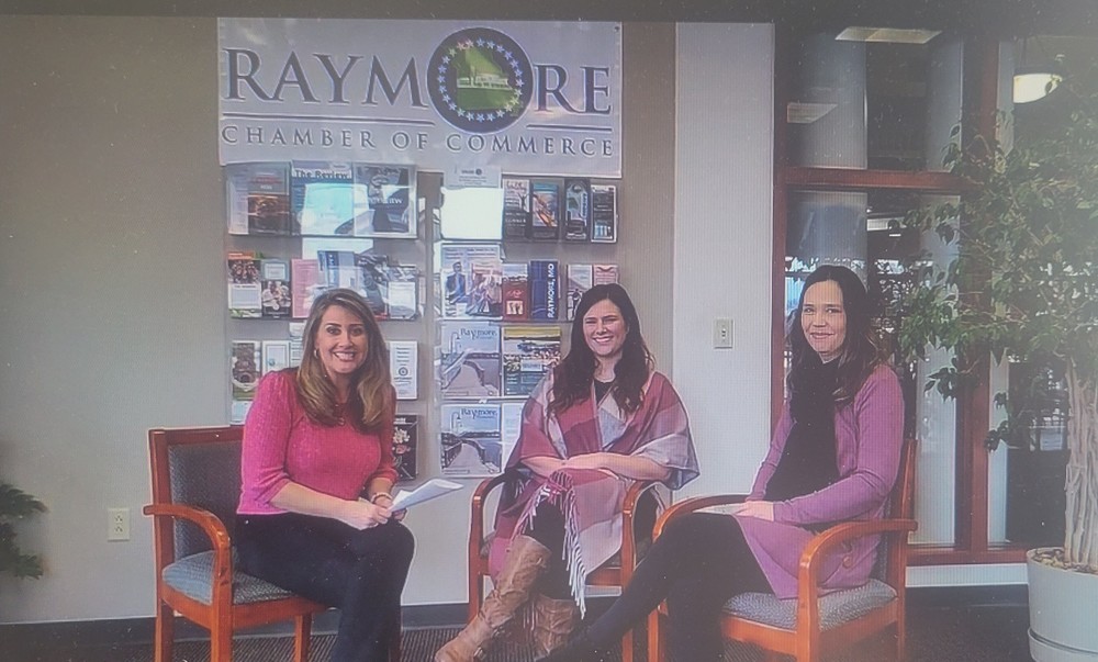 Three women sitting in chairs in front of the Raymore Chamber of Commerce sign.